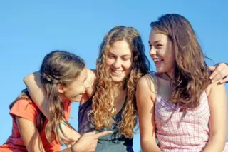 três adolescentes sorrindo sentadas juntas ao ar livre com céu azul ao fundo