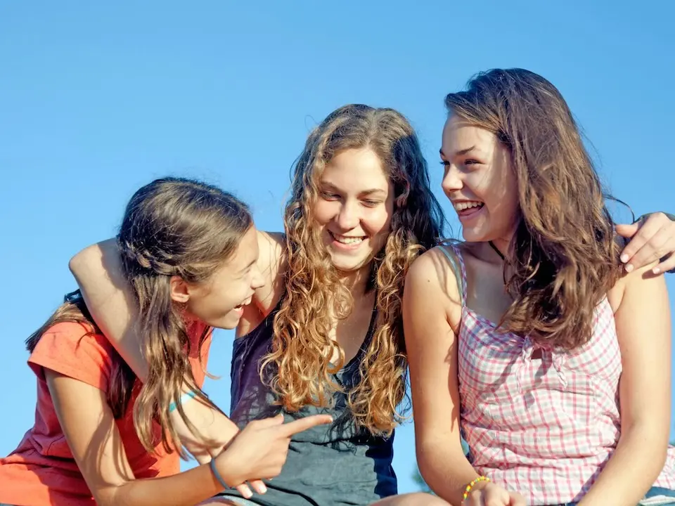 três adolescentes sorrindo sentadas juntas ao ar livre com céu azul ao fundo