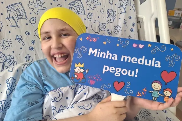 Criança em leito hospitalar com lenço amarelo na cabeça sorrindo e segurando placa azul com texto "Minha medula pegou!"