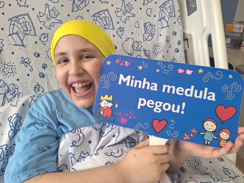 Criança em leito hospitalar com lenço amarelo na cabeça sorrindo e segurando placa azul com texto "Minha medula pegou!"