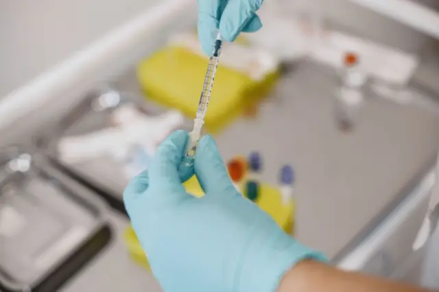 Mãos com luvas azuis segurando uma seringa sobre bandeja com materiais médicos