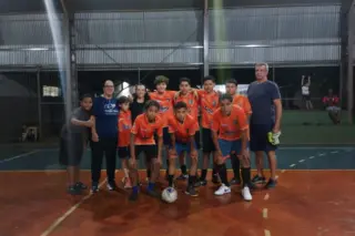 Time juvenil de futsal posando em quadra coberta; jogadores com uniformes laranja, bola à frente e dois adultos ao lado