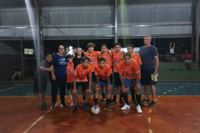Time juvenil de futsal posando em quadra coberta; jogadores com uniformes laranja, bola à frente e dois adultos ao lado