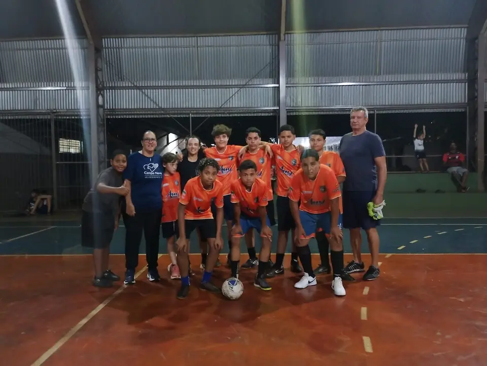 Time juvenil de futsal posando em quadra coberta; jogadores com uniformes laranja, bola à frente e dois adultos ao lado