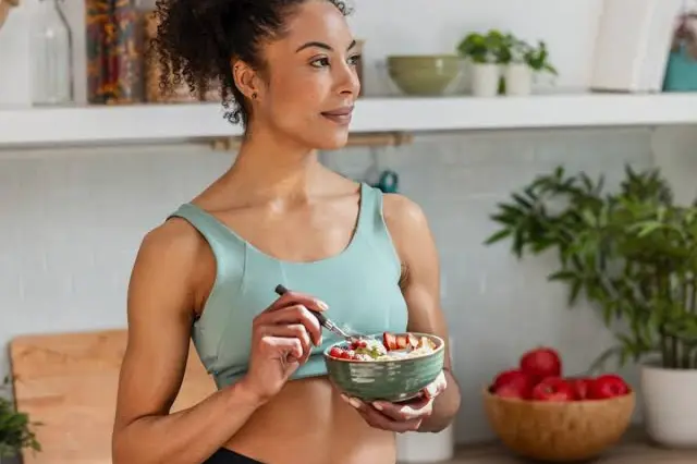 mulher jovem em uma cozinha segurando uma tigela com comida e comendo com uma colher
