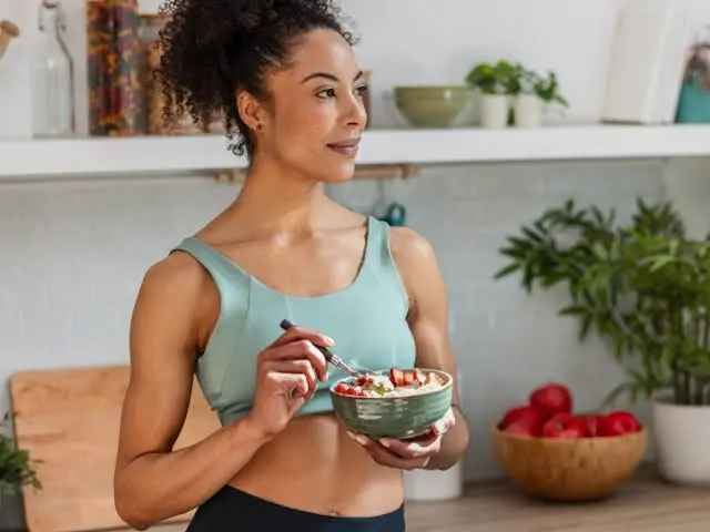 mulher jovem em uma cozinha segurando uma tigela com comida e comendo com uma colher