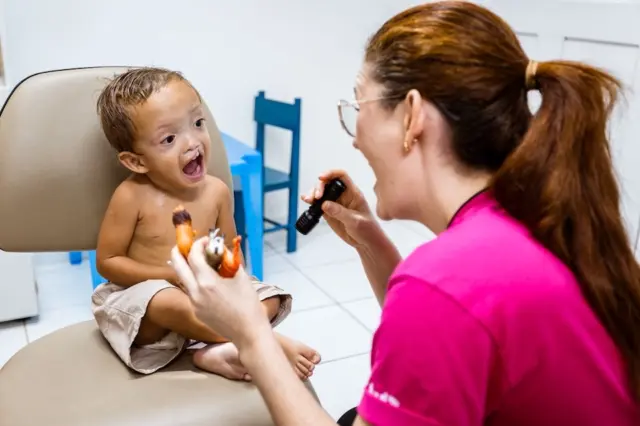 criança pequena com fissura no lábio sentada em cadeira durante consulta com profissional segurando lanterna e brinquedos