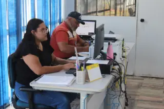 Duas pessoas sentadas em mesas de escritório trabalhando em computadores, com porta-canetas, calendário e garrafa rosa sobre a mesa
