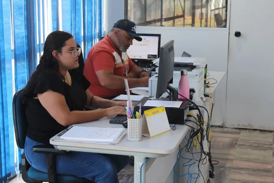 Duas pessoas sentadas em mesas de escritório trabalhando em computadores, com porta-canetas, calendário e garrafa rosa sobre a mesa
