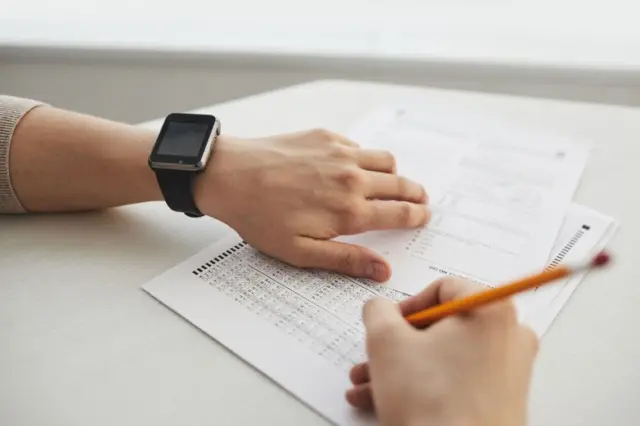 Mãos preenchendo gabarito de prova com lápis; mão esquerda apoiada com relógio smartwatch no pulso