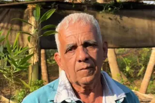 Homem idoso em close, cabelos curtos e brancos, vestindo camisa azul com gola branca, ao ar livre com vegetação ao fundo