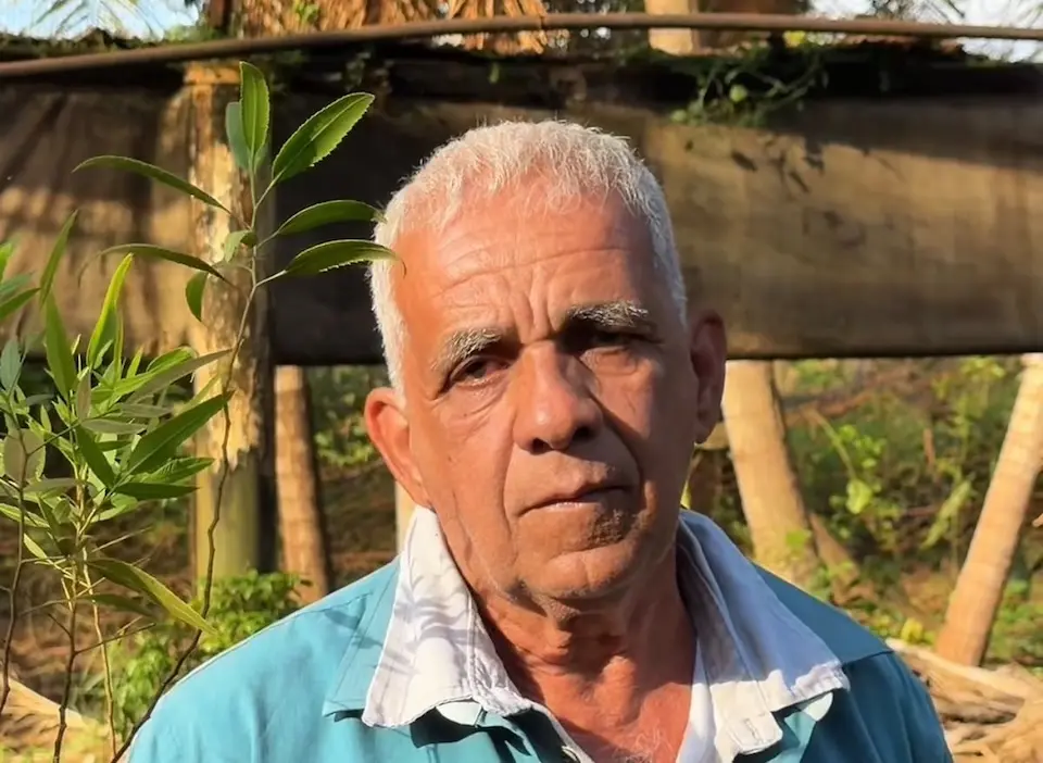 Homem idoso em close, cabelos curtos e brancos, vestindo camisa azul com gola branca, ao ar livre com vegetação ao fundo