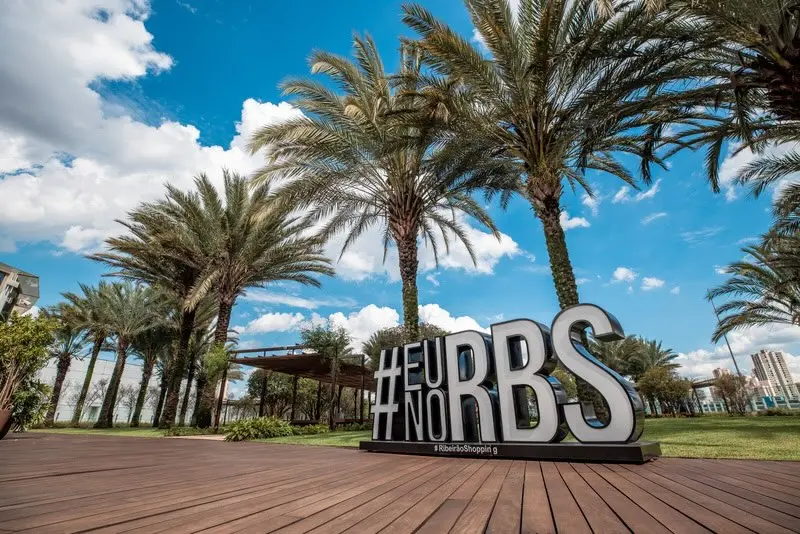 Placa em letras tridimensionais brancas em pedestal sobre deck de madeira, palmeiras e céu azul ao fundo