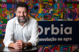 homem adulto de barba e cabelo escuros, camisa branca, sorrindo e apoiado com os antebraços sobre uma mesa