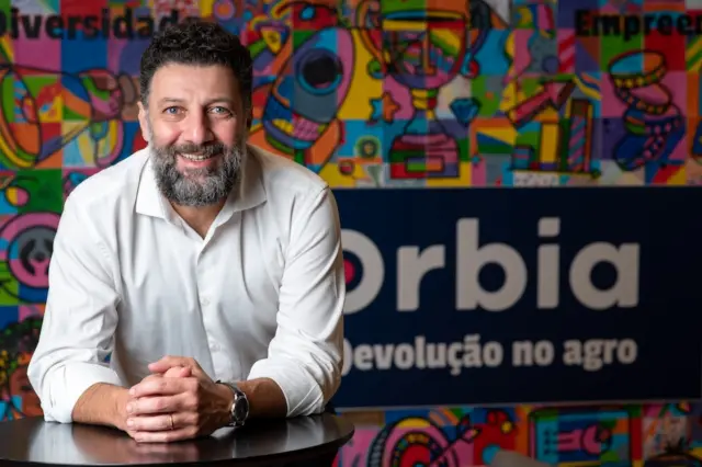 homem adulto de barba e cabelo escuros, camisa branca, sorrindo e apoiado com os antebraços sobre uma mesa