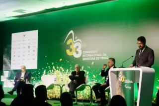 Palco com fundo verde e logotipo do 33º Congresso Internacional da Indústria do Trigo, três pessoas sentadas e um palestrante em pé no púlpito, com público em silhueta em primeiro plano