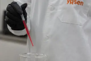 Pessoa com jaleco branco com logotipo Yosen e luva preta segurando pipeta com líquido vermelho sobre um béquer com líquido transparente