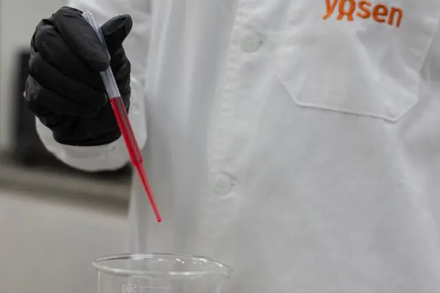 Pessoa com jaleco branco com logotipo Yosen e luva preta segurando pipeta com líquido vermelho sobre um béquer com líquido transparente