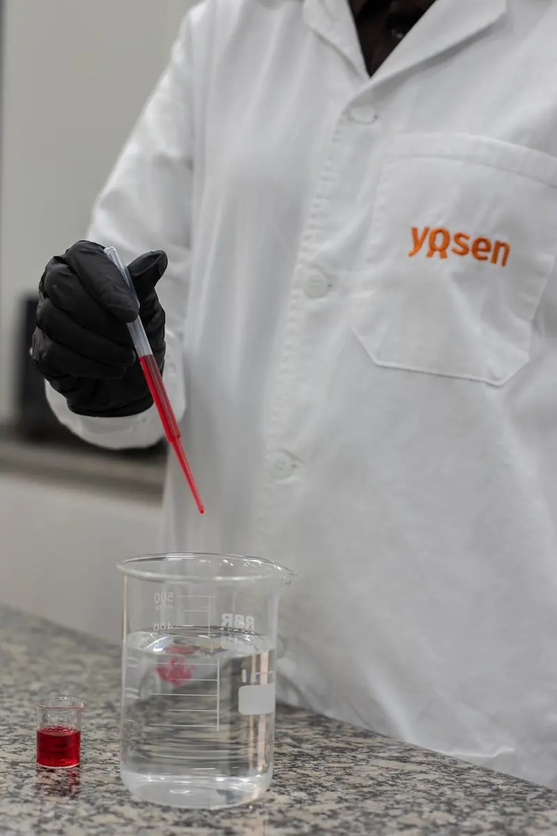 Pessoa com jaleco branco com logotipo Yosen e luva preta segurando pipeta com líquido vermelho sobre um béquer com líquido transparente