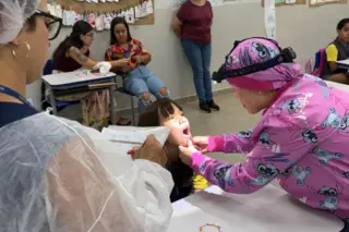 Profissional com roupa rosa e lanterna de cabeça examinando a criança com a boca aberta em sala de aula; outra funcionária de branco observa segurando papéis.