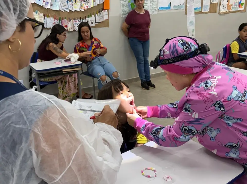 Profissional com roupa rosa e lanterna de cabeça examinando a criança com a boca aberta em sala de aula; outra funcionária de branco observa segurando papéis.