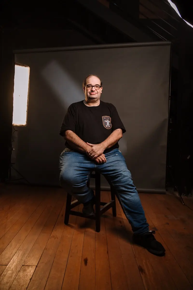homem sentado em banqueta diante de fundo cinza em estúdio fotográfico, com softboxes à esquerda e à direita