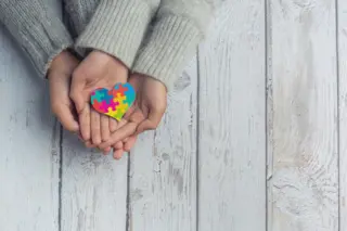 mãos de adulto e de criança apoiadas com um coração de peças de quebra-cabeça coloridas sobre tábuas de madeira claras