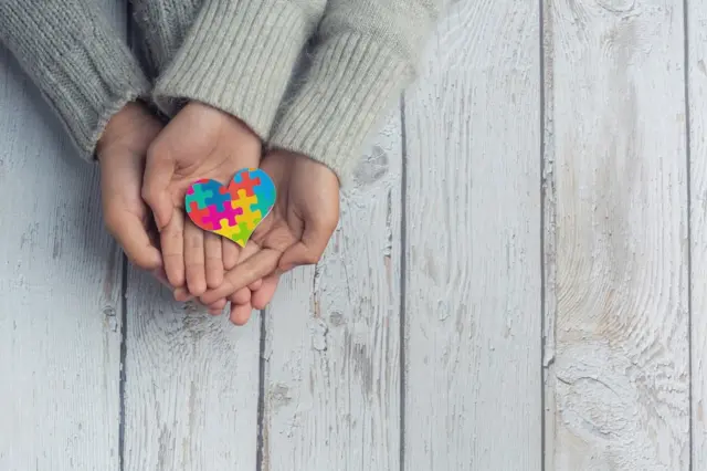 mãos de adulto e de criança apoiadas com um coração de peças de quebra-cabeça coloridas sobre tábuas de madeira claras