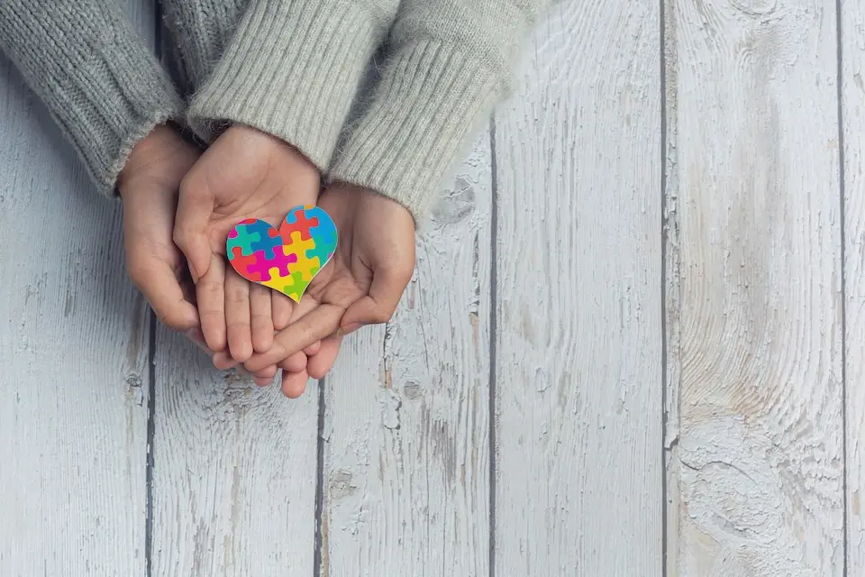 mãos de adulto e de criança apoiadas com um coração de peças de quebra-cabeça coloridas sobre tábuas de madeira claras