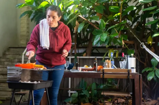 Mulher prepara alimentos em churrasqueira portátil ao lado de mesa com bebidas e utensílios em área externa com plantas.