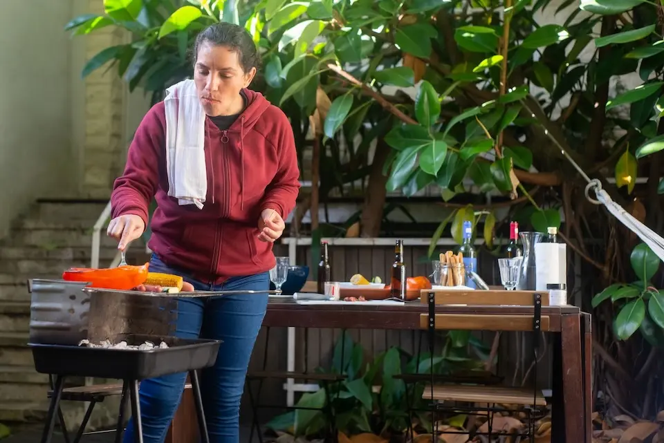 Mulher prepara alimentos em churrasqueira portátil ao lado de mesa com bebidas e utensílios em área externa com plantas.