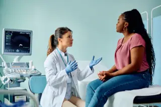 Médica de jaleco e luvas conversando com paciente sentada em maca, com monitor de ultrassom ao fundo
