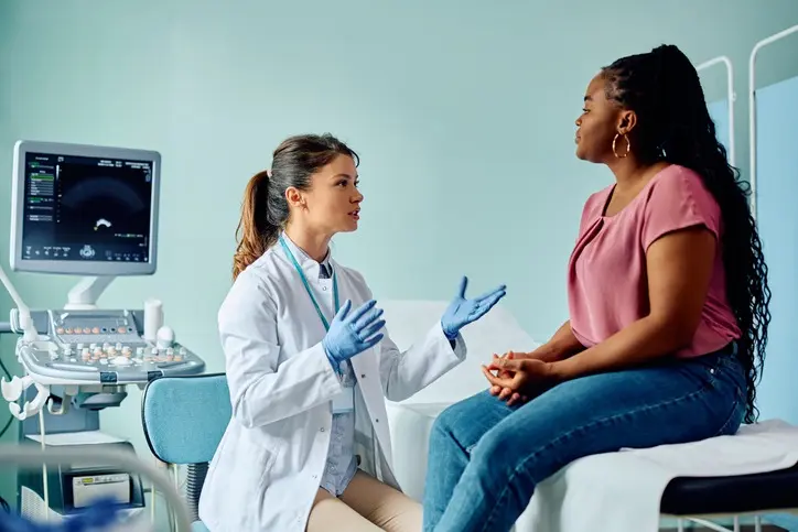 Médica de jaleco e luvas conversando com paciente sentada em maca, com monitor de ultrassom ao fundo