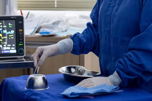 Mãos de profissional com avental cirúrgico azul e luvas brancas manuseando pinça sobre campo azul com tigelas e bandejas de aço inox; monitor médico ao fundo
