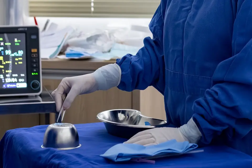 Mãos de profissional com avental cirúrgico azul e luvas brancas manuseando pinça sobre campo azul com tigelas e bandejas de aço inox; monitor médico ao fundo