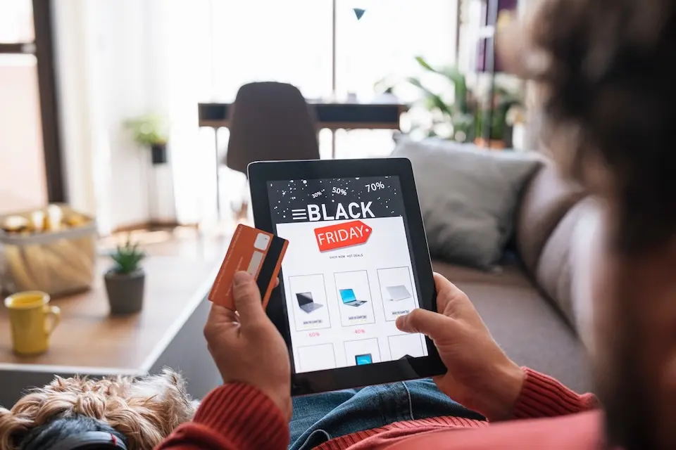 Pessoa segurando tablet que mostra anúncio 'BLACK FRIDAY' e cartão de crédito, sentada em sofá com cachorro ao lado