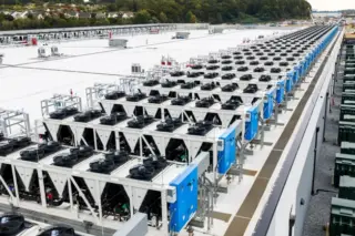 Fileiras de chillers industriais brancos e azuis com ventiladores visíveis no telhado de um data center, com tubulações e área verde ao fundo