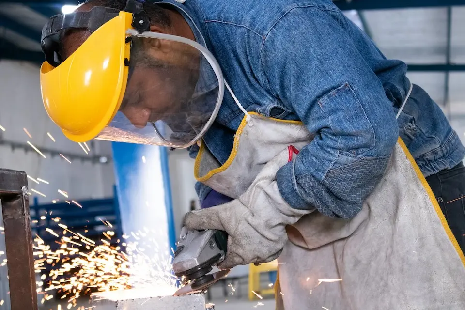 Trabalhador usando capacete amarelo com viseira, luvas e avental enquanto opera uma esmerilhadeira que gera faíscas