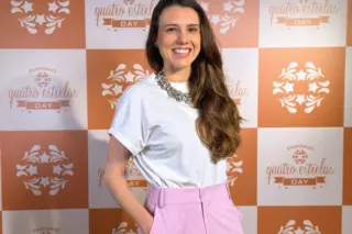 mulher adulta sorrindo em pé com as mãos nos bolsos, vestindo camiseta branca, colar grande e calça rosa, diante de painel quadriculado laranja e branco com o logotipo 'quatro estrelas' repetido