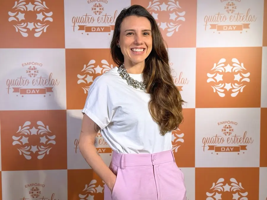 mulher adulta sorrindo em pé com as mãos nos bolsos, vestindo camiseta branca, colar grande e calça rosa, diante de painel quadriculado laranja e branco com o logotipo 'quatro estrelas' repetido