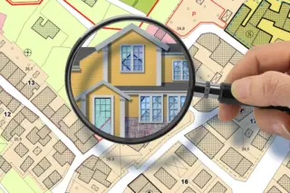 mão segurando lupa que amplia ilustração de uma casa amarela sobre mapa cadastral colorido com lotes