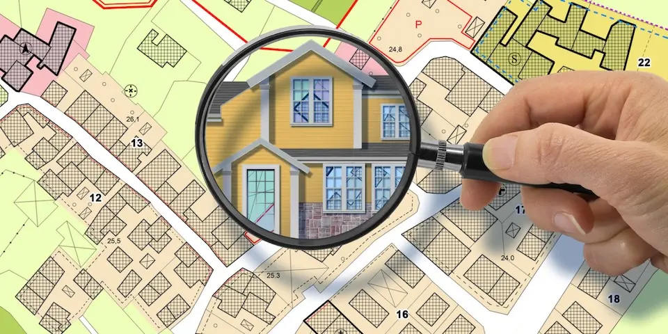 mão segurando lupa que amplia ilustração de uma casa amarela sobre mapa cadastral colorido com lotes