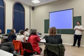 Pessoas sentadas em sala de aula assistindo a um homem em pé ao lado de uma tela de projeção em branco