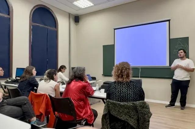 Pessoas sentadas em sala de aula assistindo a um homem em pé ao lado de uma tela de projeção em branco