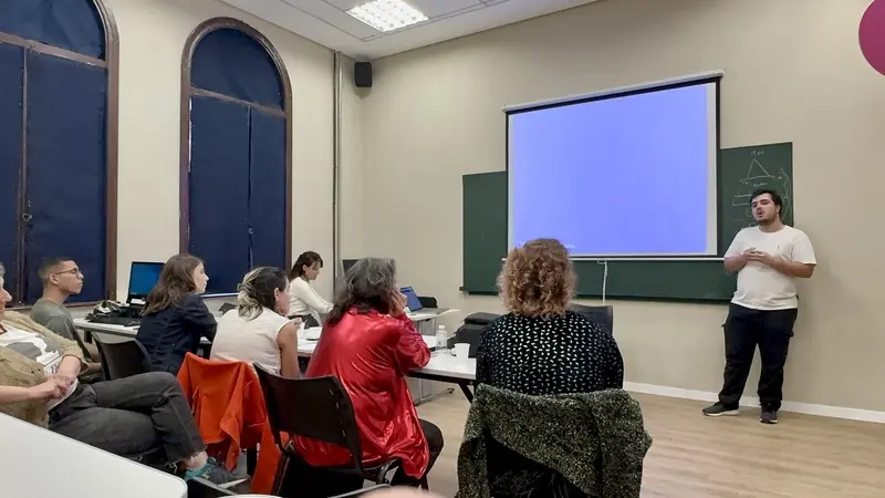 Pessoas sentadas em sala de aula assistindo a um homem em pé ao lado de uma tela de projeção em branco