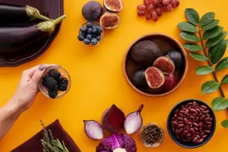 Fundo amarelo com disposição de frutas, vegetais e leguminosas: figos cortados, abacates, uvas, mirtilos, berinjela, repolho roxo, cebola roxa, beterraba, feijão e uma mão segurando uma tigela com amoras.