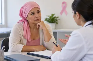 Mulher adulta com lenço rosa na cabeça sentada à mesa de consultório conversando com profissional de saúde; laço rosa afixado na parede ao fundo