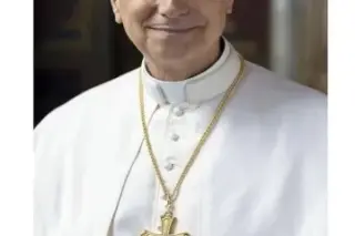 Homem idoso com batina branca e crucifixo dourado ao peito, sorrindo levemente, fundo interno desfocado