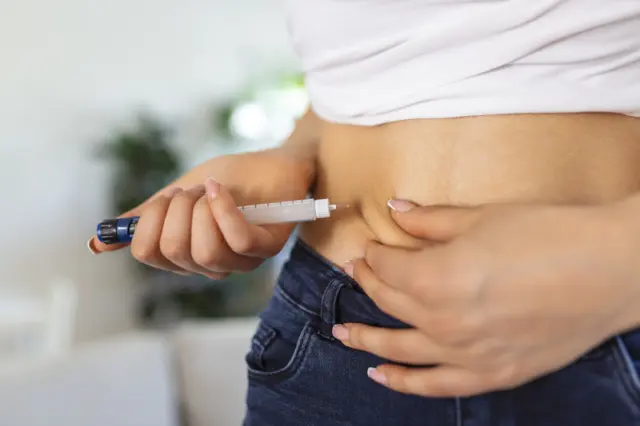 mãos segurando caneta de injeção perto do abdômen, com dobra de pele visível; pessoa usa calça jeans e blusa branca