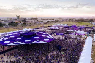 Vista aérea de festival ao ar livre ao entardecer com grande público, palco com cobertura roxa em formato geométrico, tendas brancas e pessoas vestindo camisetas roxas